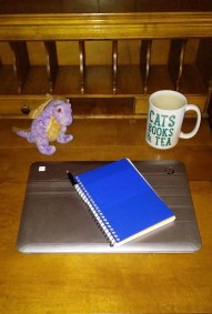 A photo of a wooden fold-out desk with a closed laptop, a blue notebook, a pen, a mug reading "cats, books, and tea" and a purple and gold dragon stuffed animal